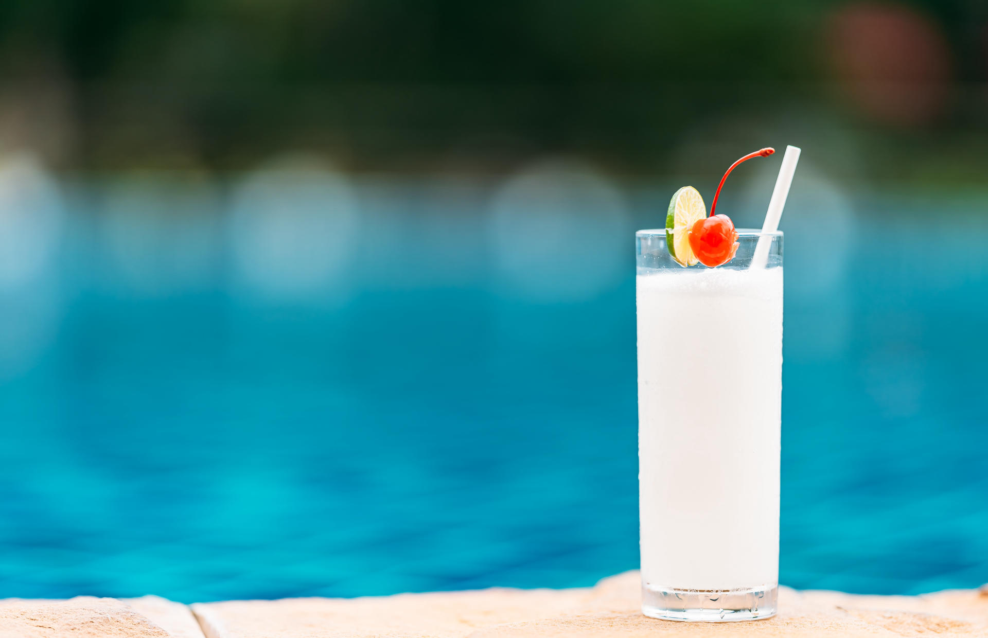 A coconut juice drink with a lime slice and a cherry sits at the edge of a pool.