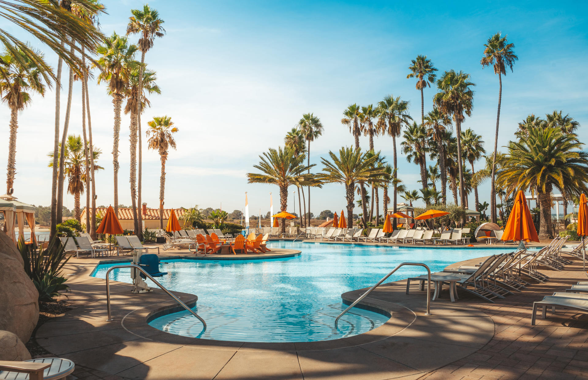 A hotel pool is surrounded by palm trees and orange umbrellas.