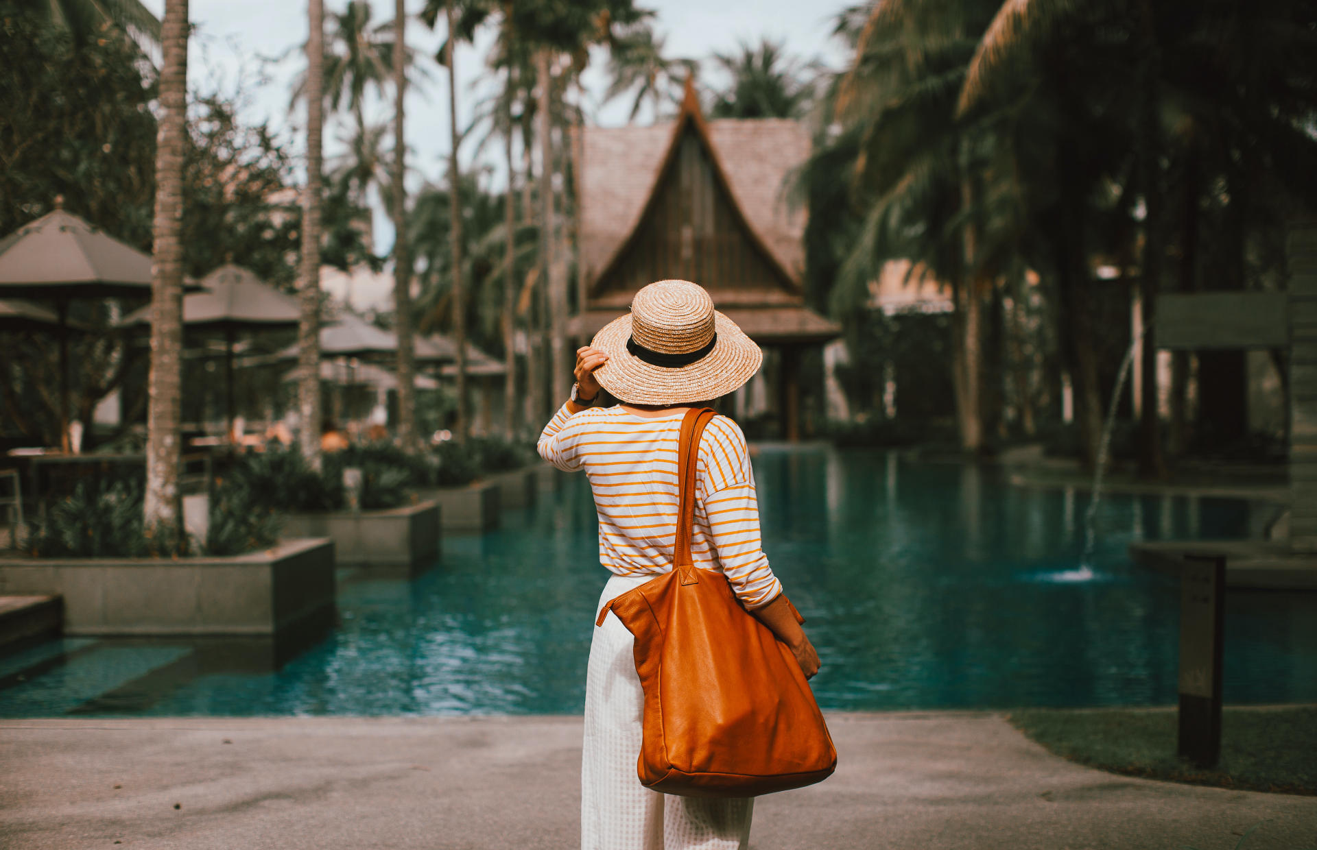 A guest wearing a sun hat and carrying at tote bag overlooks a hotel pool.