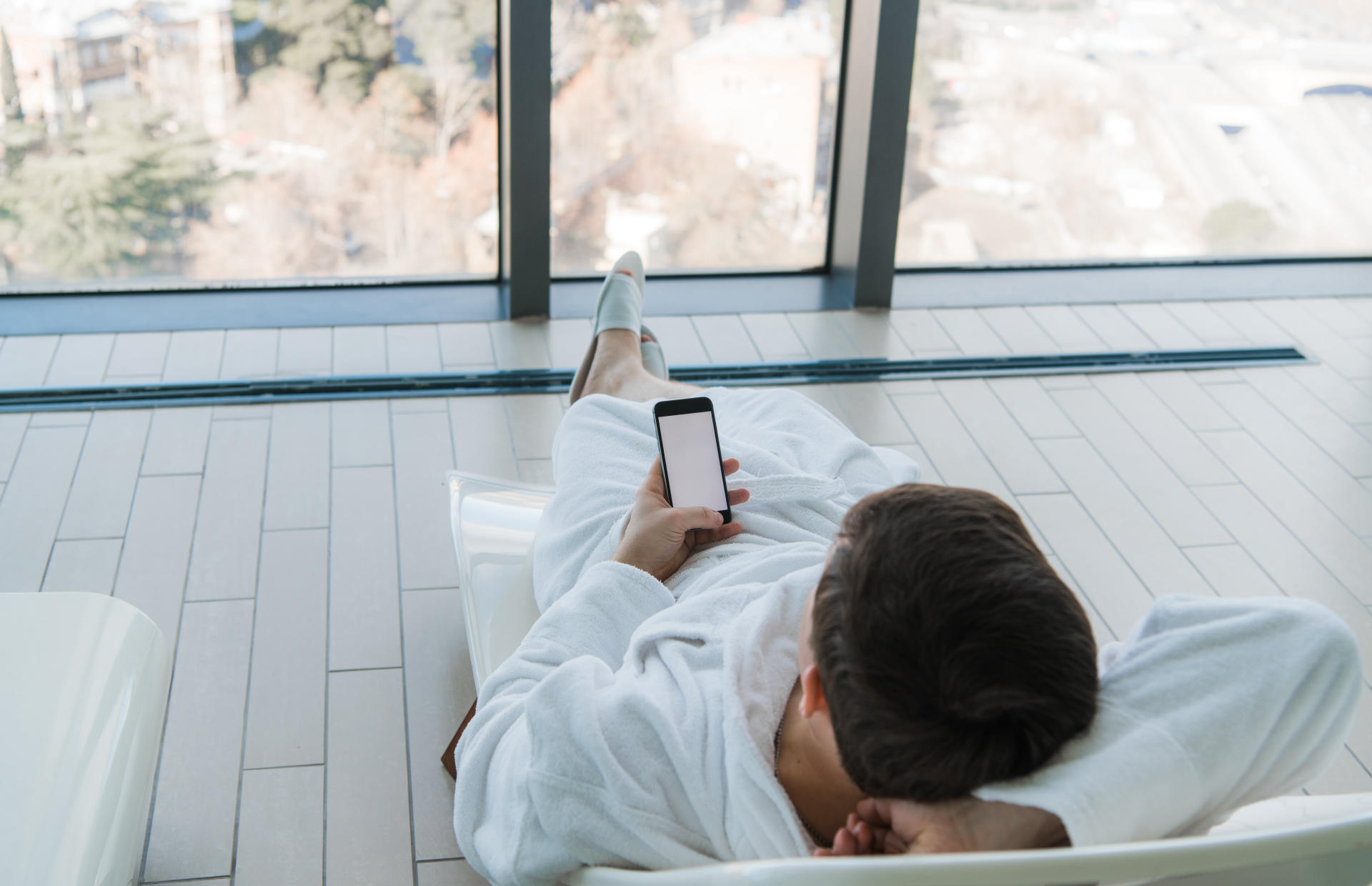 A man in a white robe reclining on a hotel chair looking at his phone.