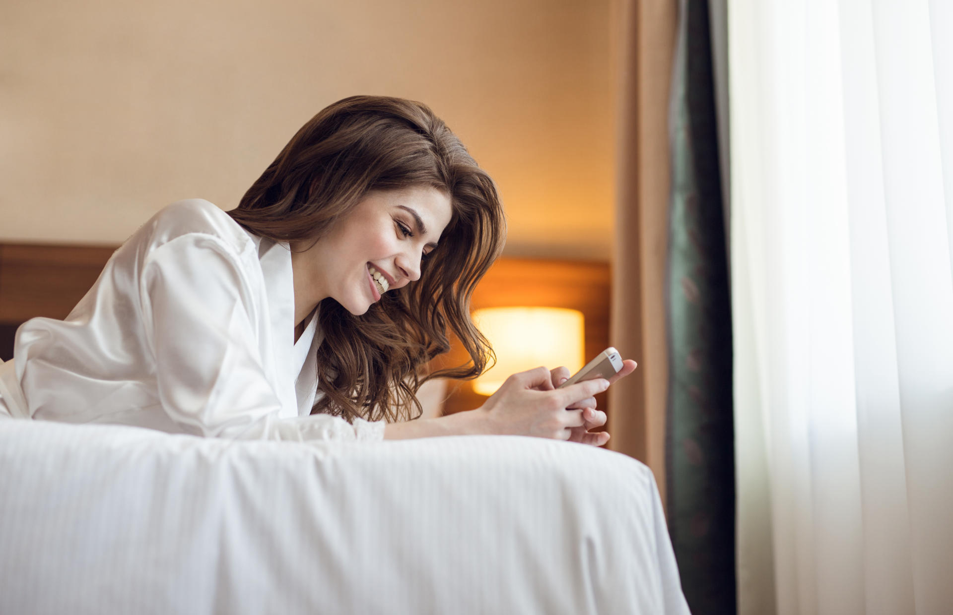 A woman is laying down on her stomach on stomach on a hotel bed looking at her phone.