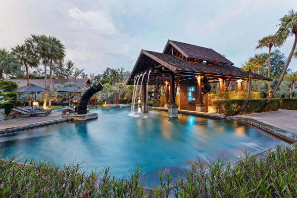 A pool with a waterfall element surrounded by chairs, umbrellas, and palm trees at The Slate, a Phuket Pearl Resort