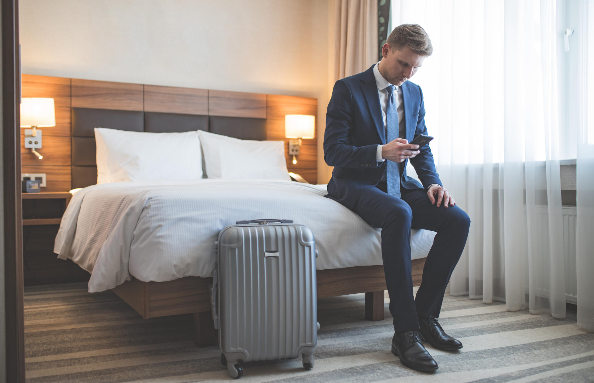 A business man is sitting on the edge of a hotel bed looking down at his phone.